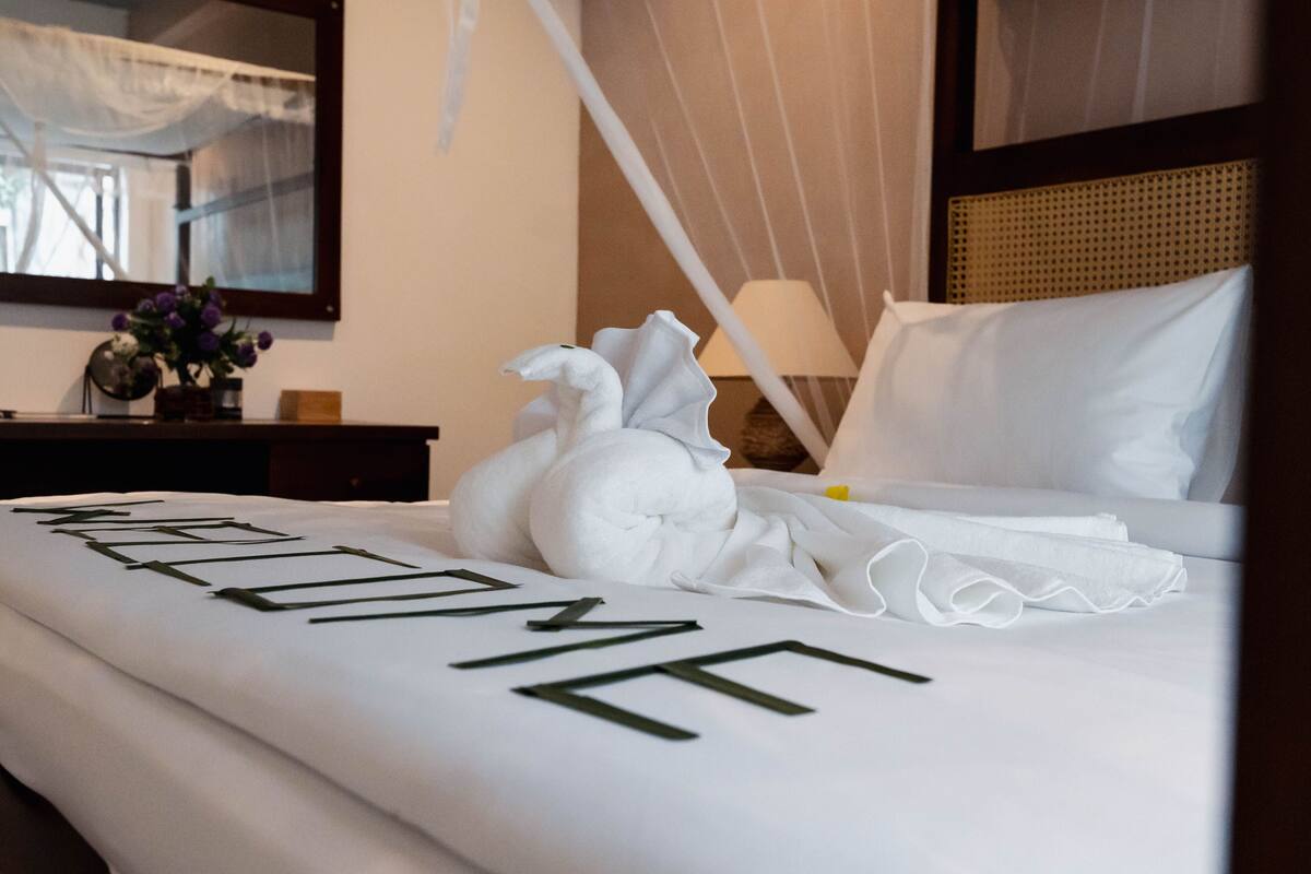 Ein Hotelzimmer mit einem ordentlich bezogenen Bett, einem Nachttisch mit Lampe, einem Spiegel und einem Fenster mit Vorhängen.