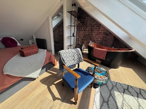 Un dormitorio tipo loft con techo inclinado, una cama con toalla enrollada, un sillón con manta estampada, un taburete pequeño y una bañera.