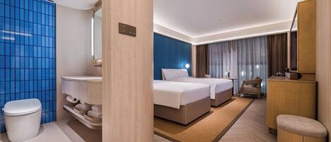 A modern hotel room with a large bed, a separate seating area, a bathroom with a toilet and sink, and a large window with curtains.