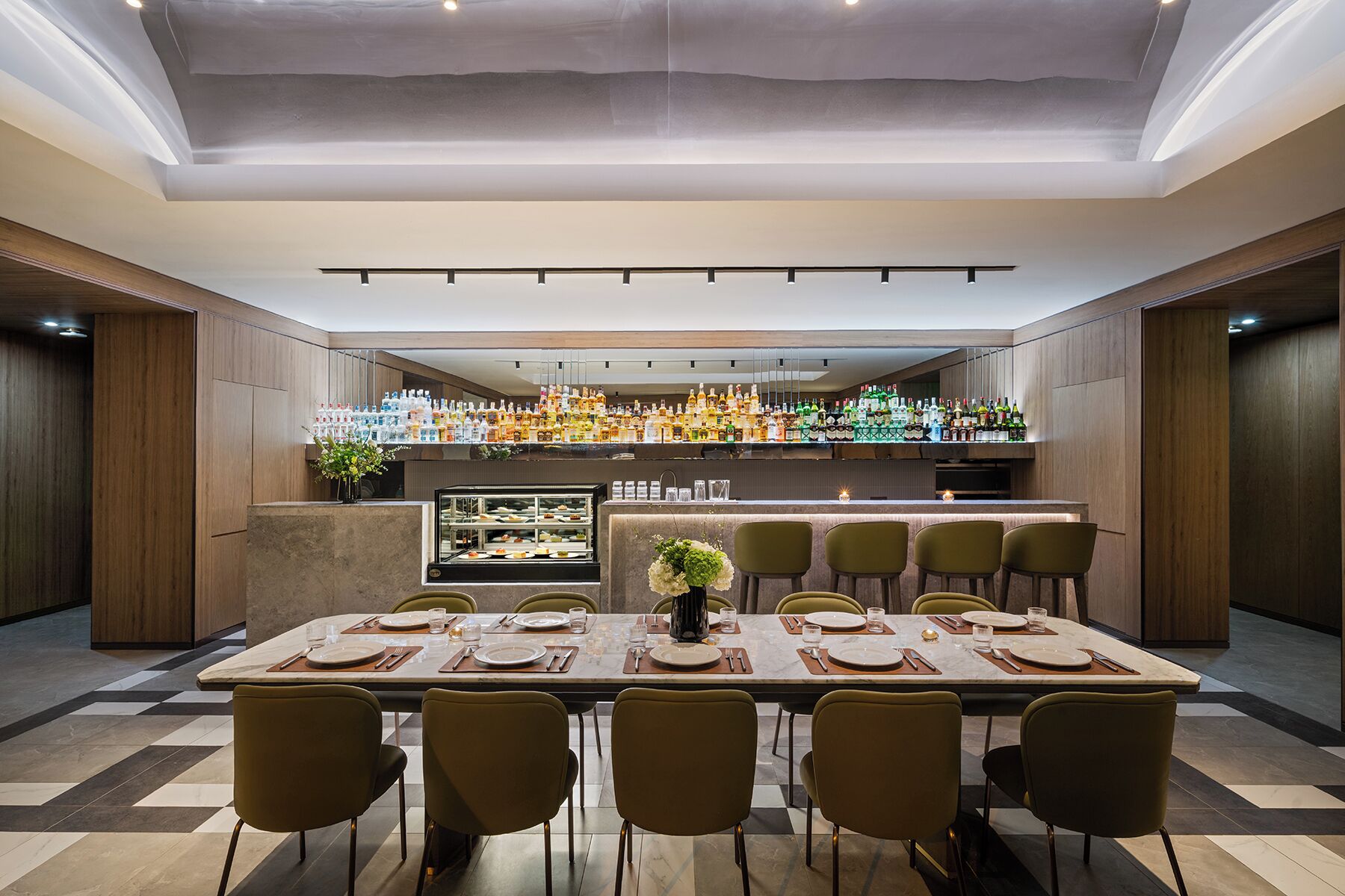 A dining area with a long table, chairs, a bar with various bottles, and a display case with food.