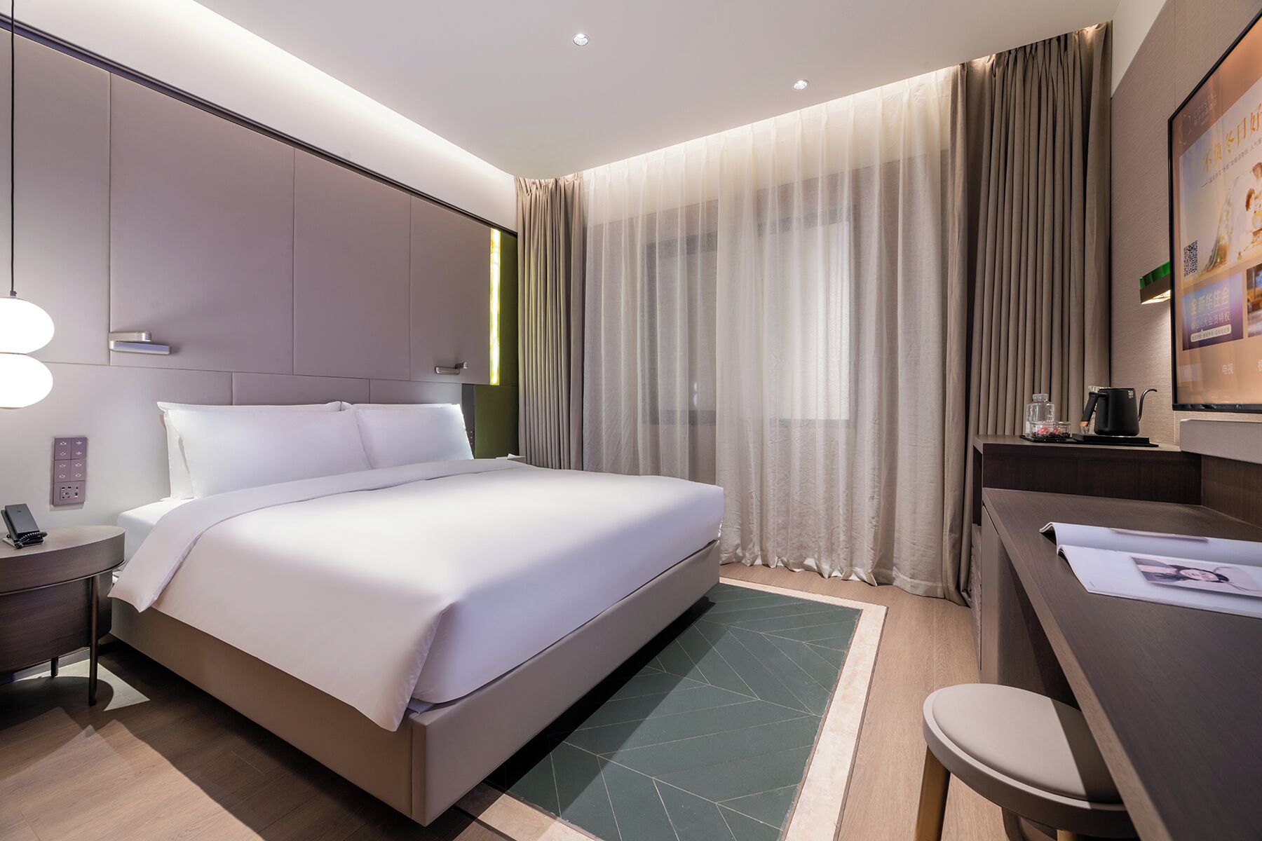 A modern hotel room with a large bed, bedside table, and a desk with a chair.