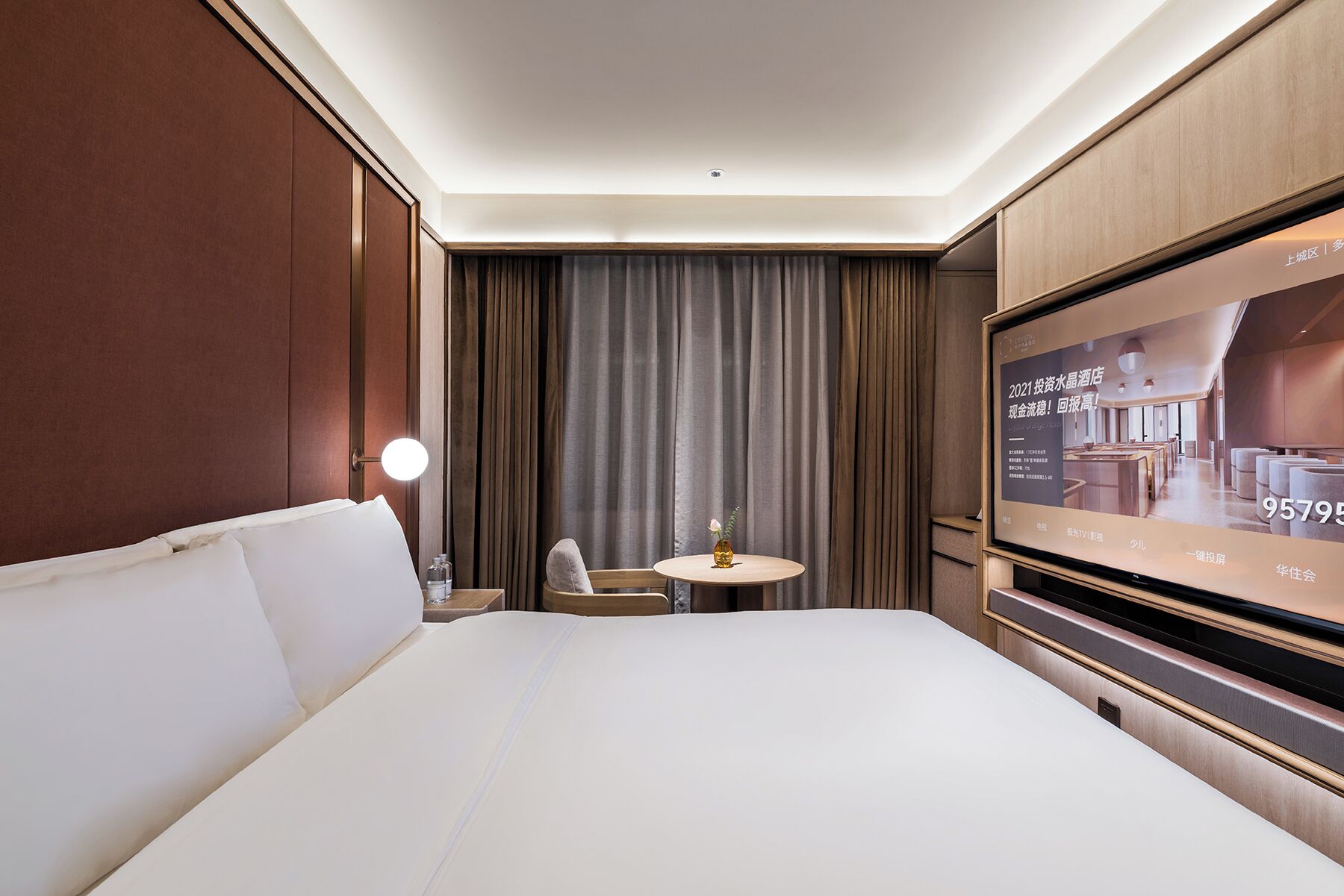 A hotel room with a large bed, a small table with a vase, a TV screen, and a curtain.