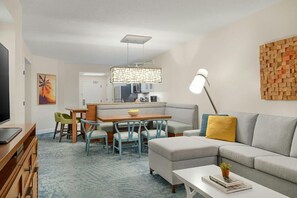 Ein modernes Wohnzimmer mit einer Couch, einem Essbereich mit Stühlen, einer Küche im Hintergrund und einem Fernseher links.
