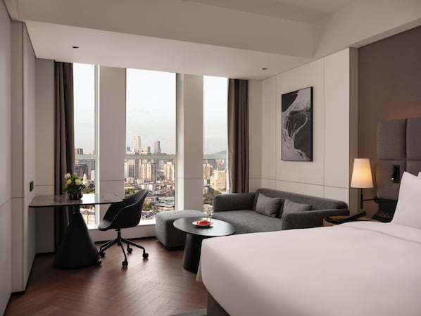 A hotel room with a bed, a sofa, a desk with a chair, a small table, and a view of a cityscape through large windows. - Intercity Hotel Qingdao Downtown (Qing Dao Shi)