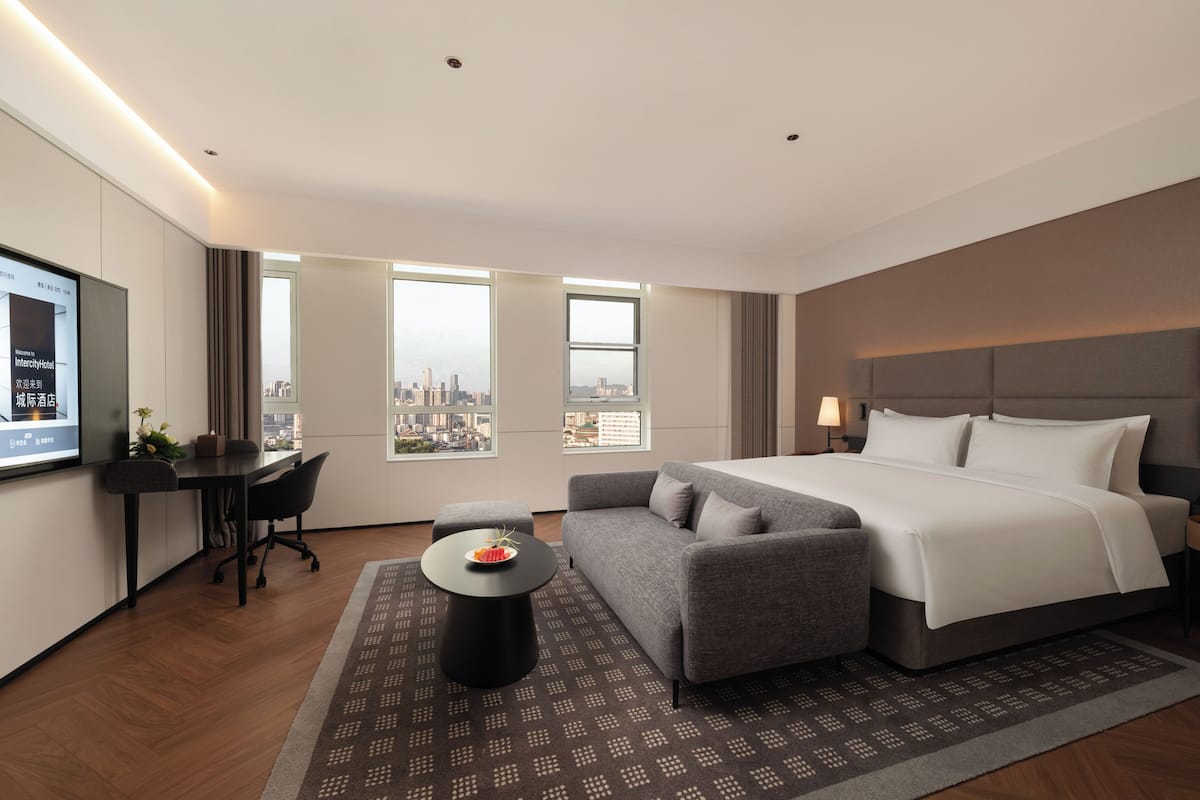 A modern hotel room with a large bed, a sofa, a desk, and a TV screen displaying a welcome message.