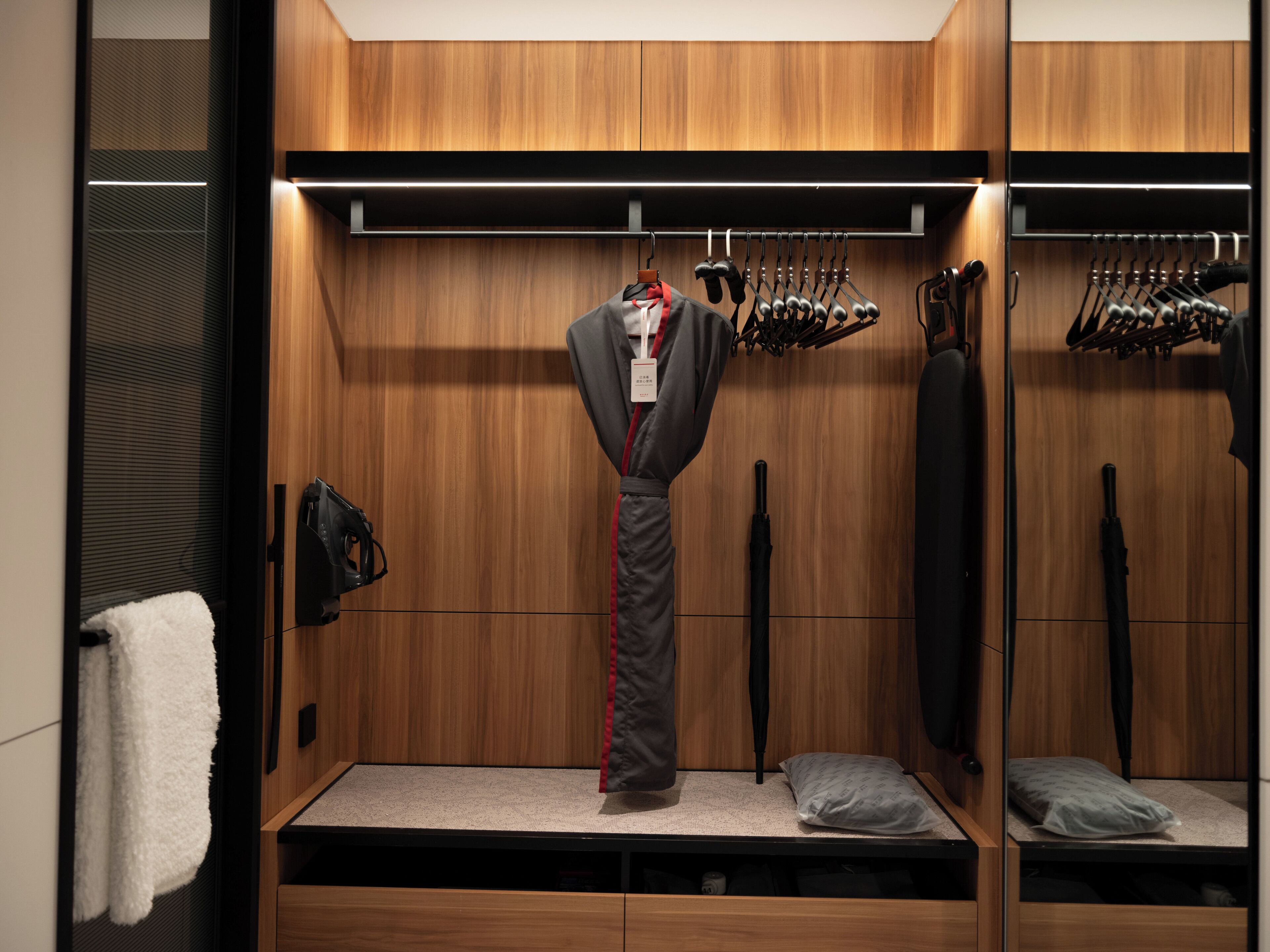 A compact, modern closet with wooden paneling, a hanging rack for shoes, a bench with a pillow, and a coat rack with a suit.