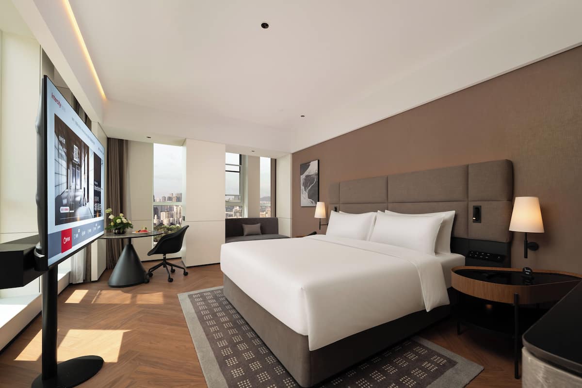 A modern hotel room with a large bed, a desk with a chair, a TV, and a city view.