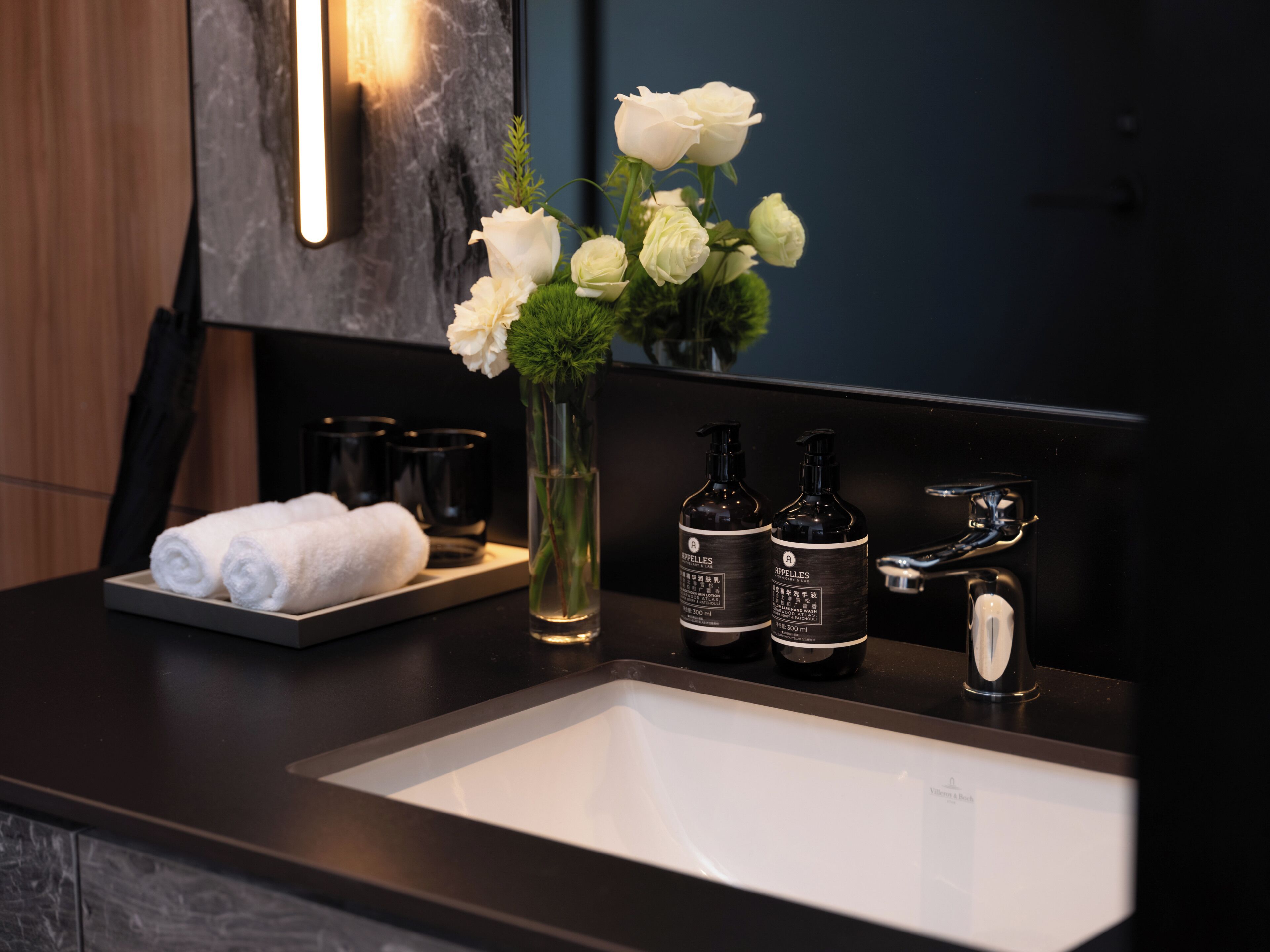A bathroom with a white sink, two bottles of hand soap, a vase with white flowers, and a towel rack with folded towels.