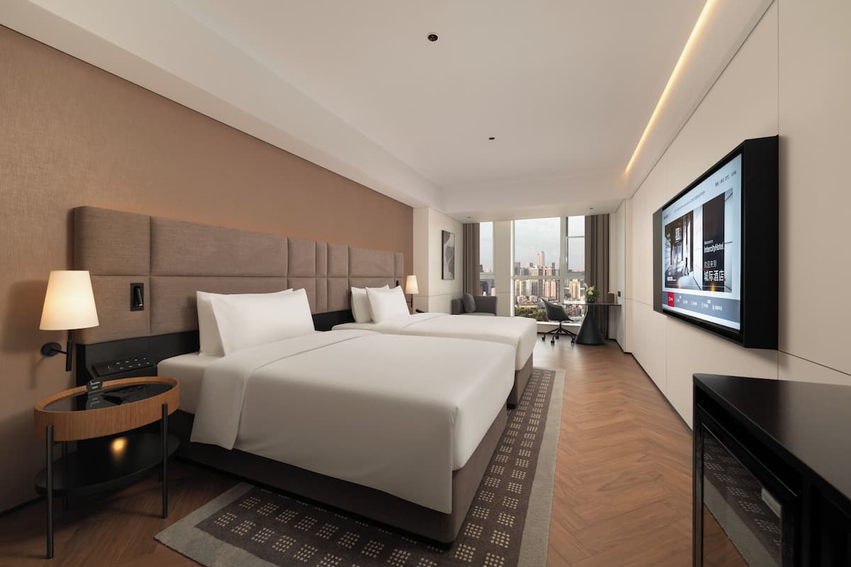 A hotel room with two beds, a TV, a desk, and a view of the city.