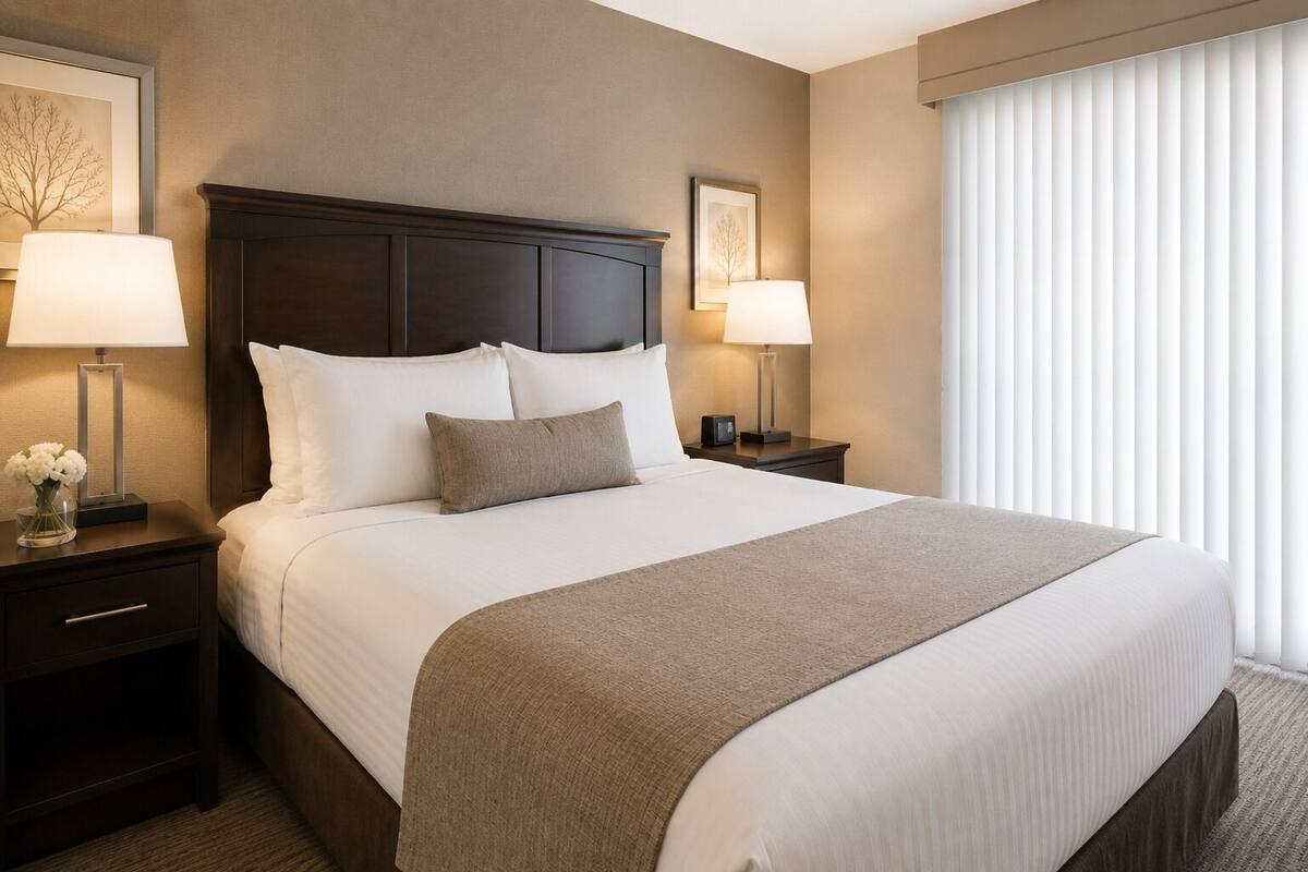 Una habitación de hotel con una cama grande, dos mesitas de noche con lámparas, un pequeño mueble y una ventana con persianas.