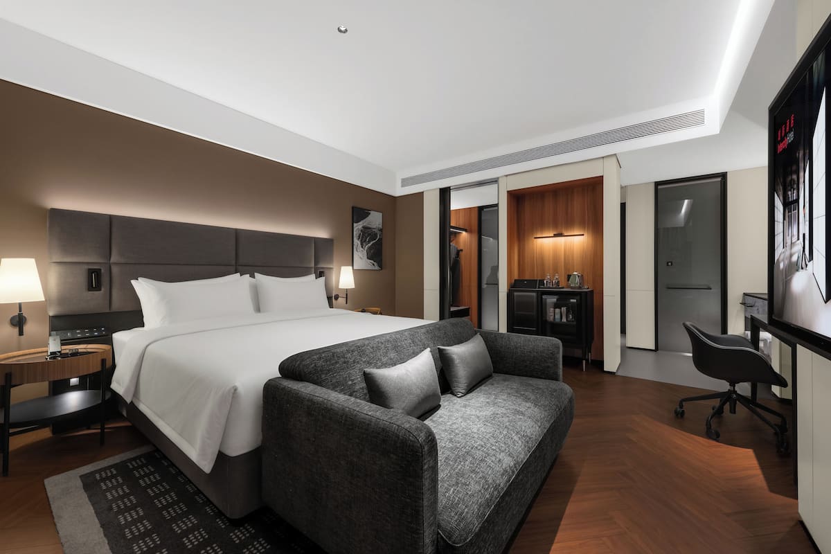 A modern hotel room with a large bed, a grey sofa, a desk, and a kitchenette.