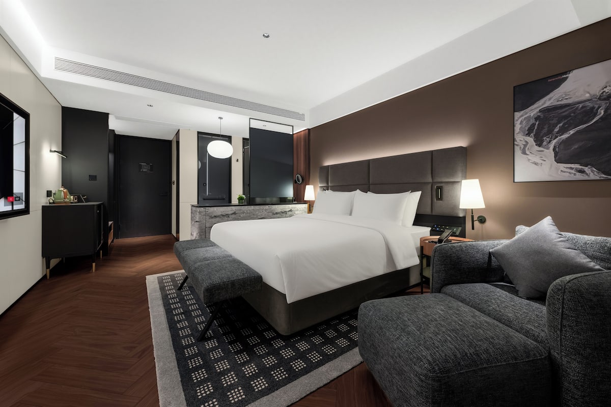 A modern hotel room with a large bed, a sofa, a desk, and a wall-mounted TV.