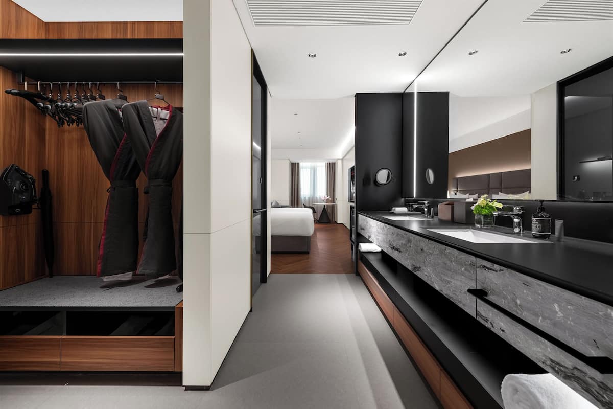 A modern hotel room with a large bed, a vanity with a mirror, and a walk-in closet with hanging clothes.