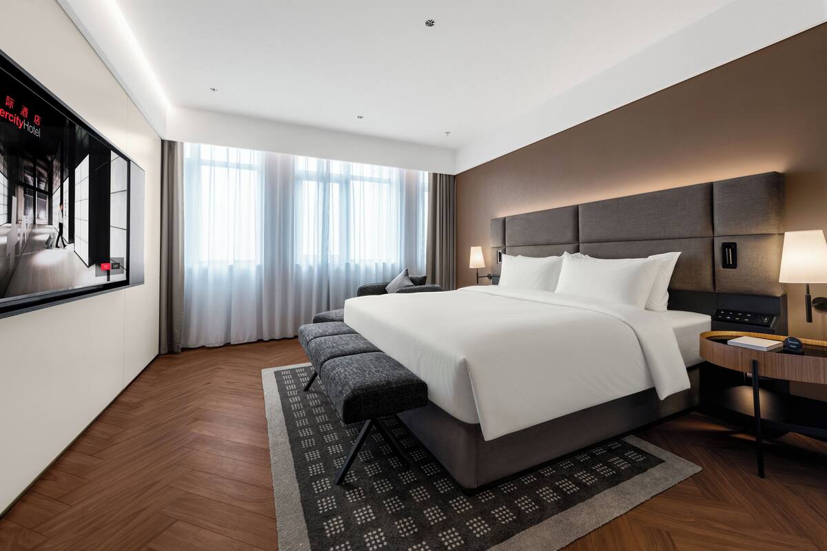 A modern hotel room with a large bed, a desk, a chair, a TV, and a large window with curtains.