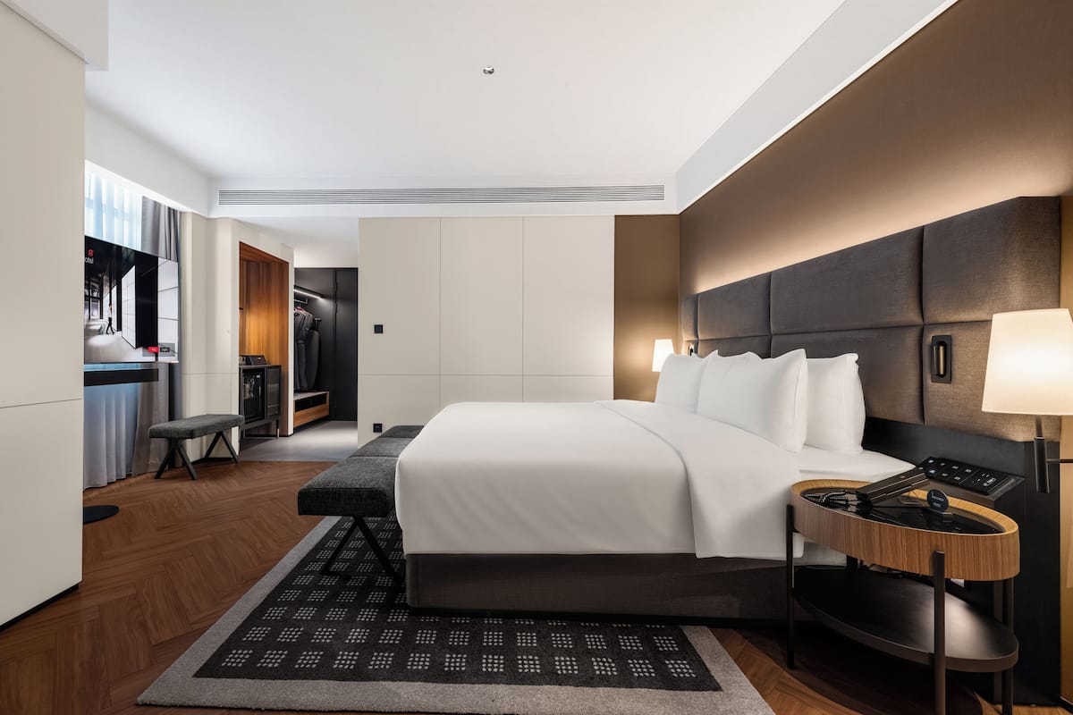 A modern hotel room with a bed, bedside table, lamp, and a view of a cityscape through the window.