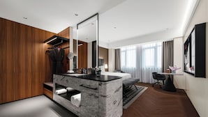 Quarto de hotel moderno com toucador de mármore, paredes revestidas a madeira, um espelho grande, uma cama com cabeceira cinzenta, uma pequena mesa com candeeiro e uma janela com cortinas.