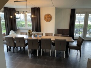 Een moderne eetruimte met een grote houten tafel, meerdere stoelen, een klok aan de muur en grote ramen met gordijnen.