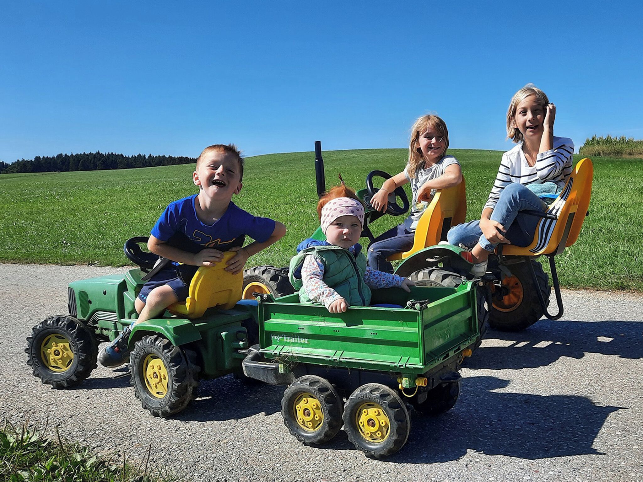Children on a green tractor with a trailer, a clear sky, and a green field in the background.