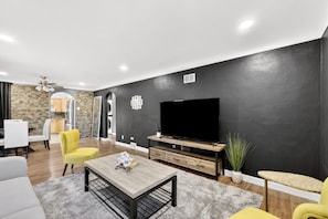 Een moderne woonkamer met een flatscreen televisie, een salontafel en een eethoek met stoelen.
