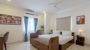 Hotelkamer met een bed, een sofa, een bureau, een stoel, een nachtkastje, een lamp, een airconditioner, een plafondventilator, een raam met gordijnen en een poster aan de muur.