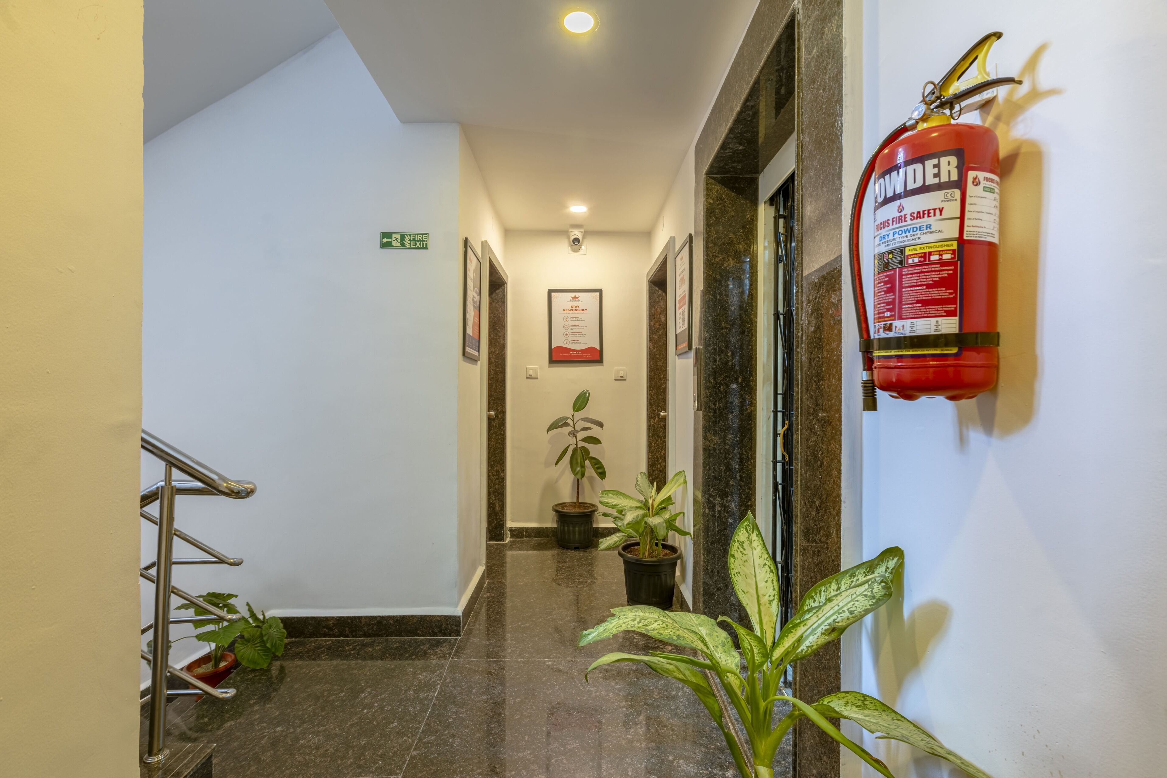 A hotel hallway with a fire extinguisher on the wall, a fire exit sign, and potted plants.