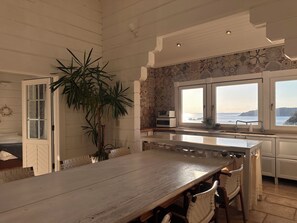 Una cocina con una gran mesa de madera, armarios blancos, una planta en maceta y vistas al mar a través de las ventanas.