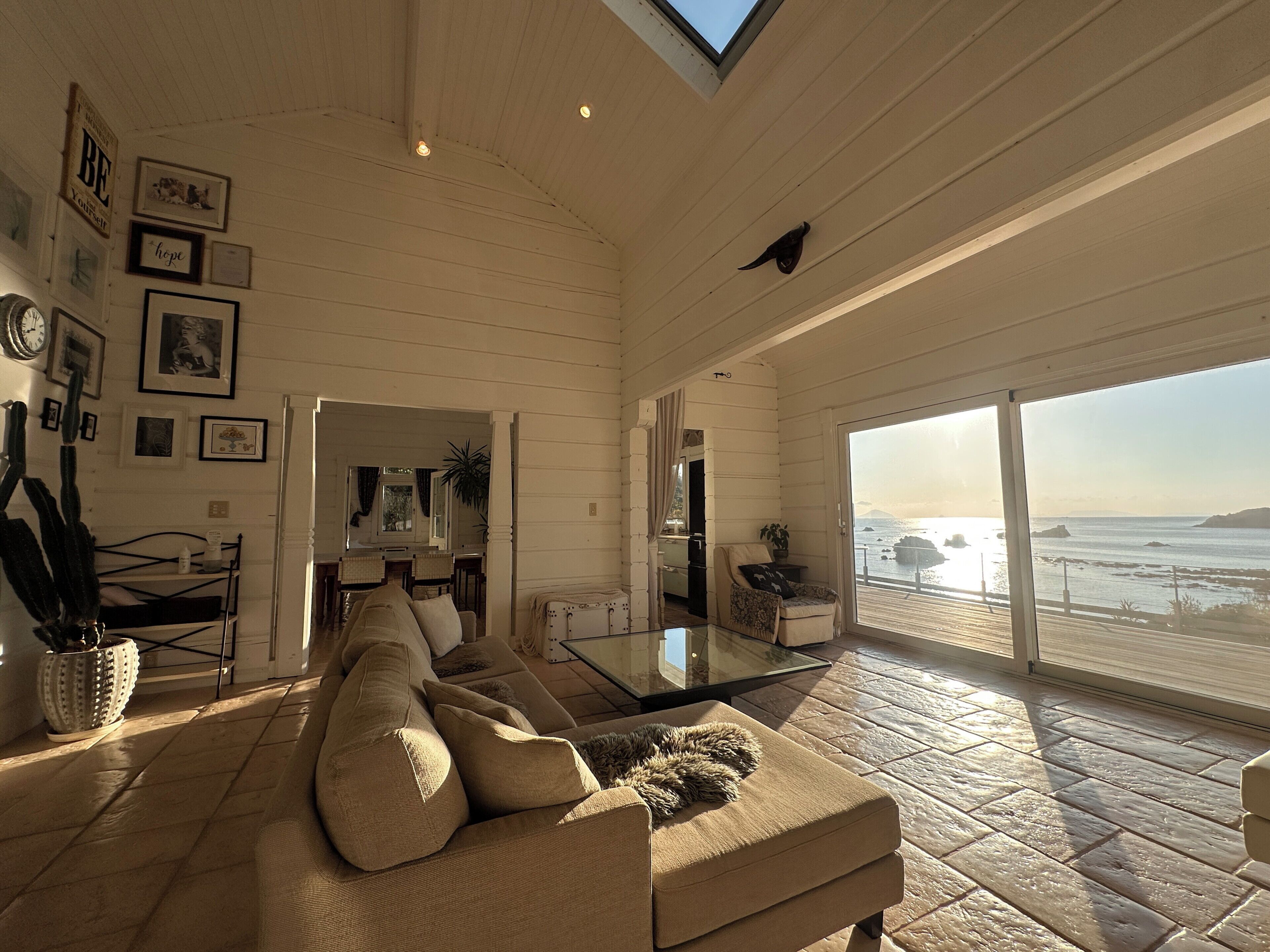 A spacious living room with a large sofa, a dining area, and a view of the sea through large windows.