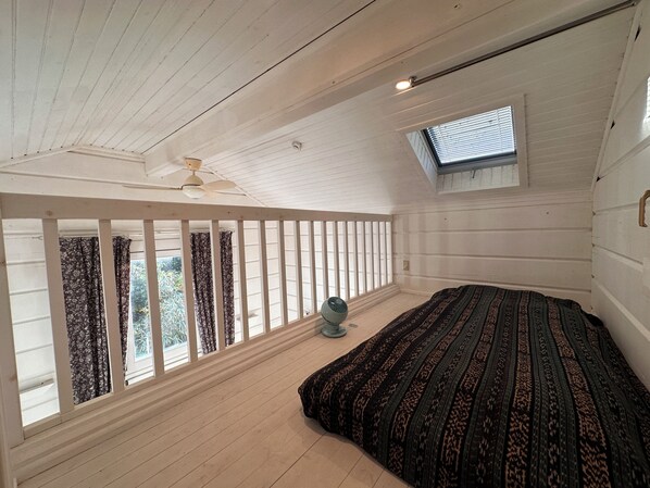 Ein Zimmer mit schrägem Dach, einem Bett mit gemustertem Überwurf, einem Standventilator und einem Dachfenster.