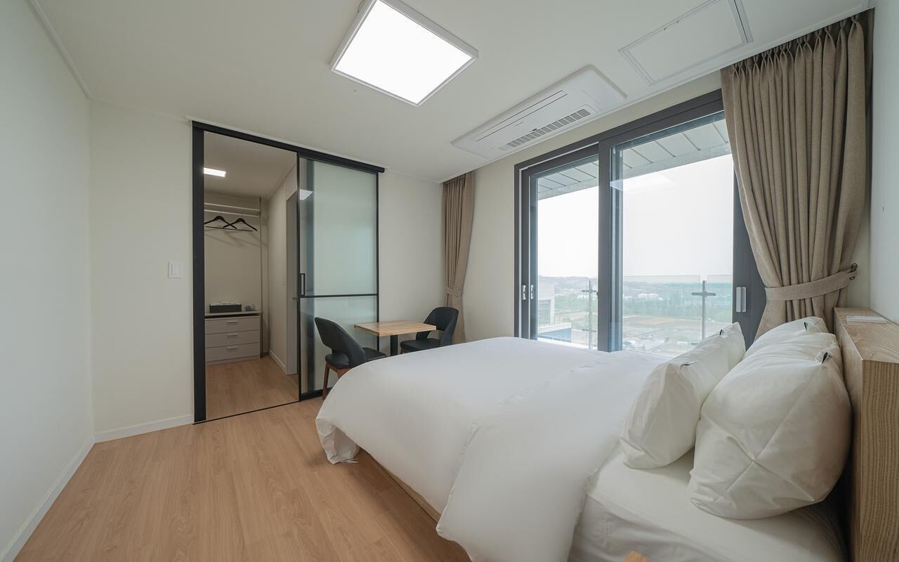 A hotel room with a large bed, a desk, and a chair, a closet, and a view of the city through a large window.