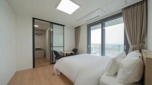 Habitación de hotel con una cama grande, un escritorio, una silla, un armario y vistas de la ciudad a través de un ventanal amplio.