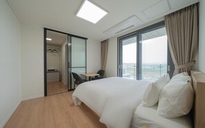 Habitación de hotel con una cama grande, un escritorio, una silla, un armario y vistas de la ciudad a través de un ventanal amplio.