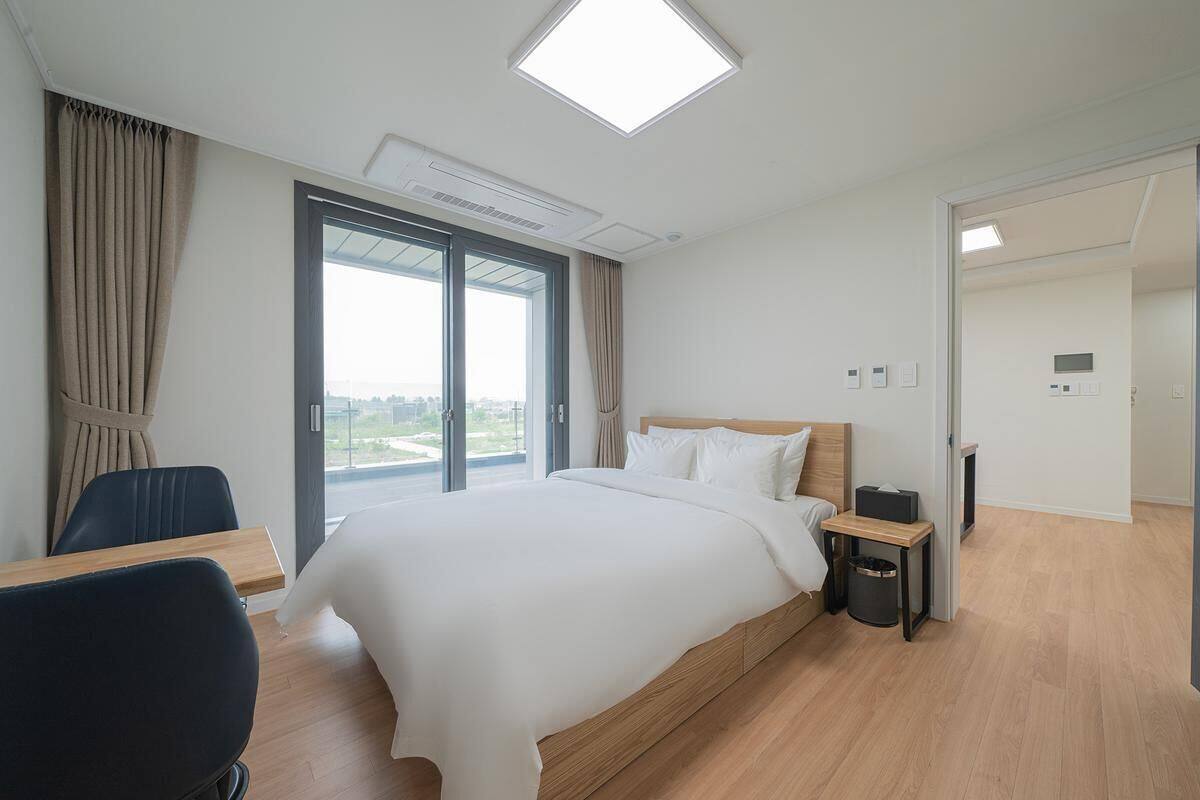 Moderni hotellihuone, jossa on suuri sänky, työpöytä, tuoli, pieni pöytä, roskakori ja näkymä suuresta ikkunasta.