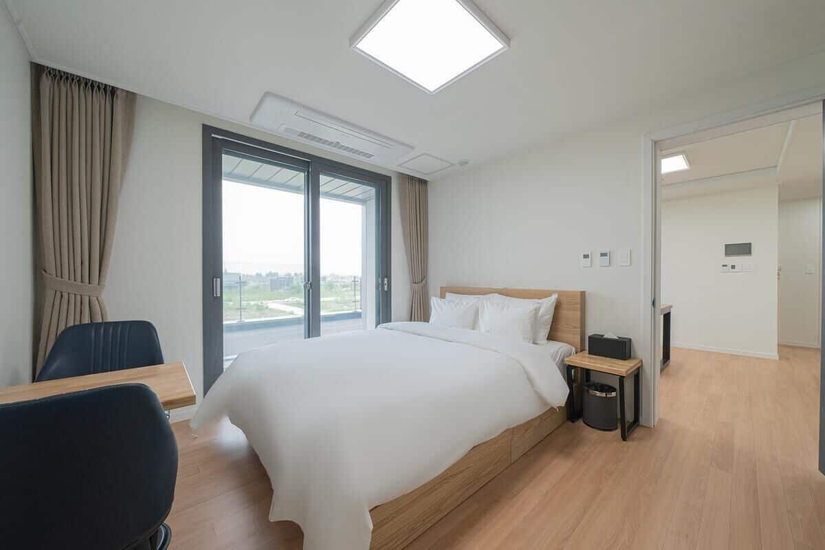A modern hotel room with a large bed, a desk, a chair, a small table, a trash bin, and a view through a large window.