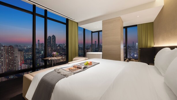 Una habitación de hotel moderna con una cama grande, una mesita con una revista y fruta, y vistas a la ciudad a través de amplios ventanales.