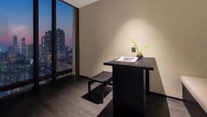 Una habitación de hotel moderna con un gran ventanal que ofrece vistas a la ciudad, un pequeño escritorio con una planta y un banco.