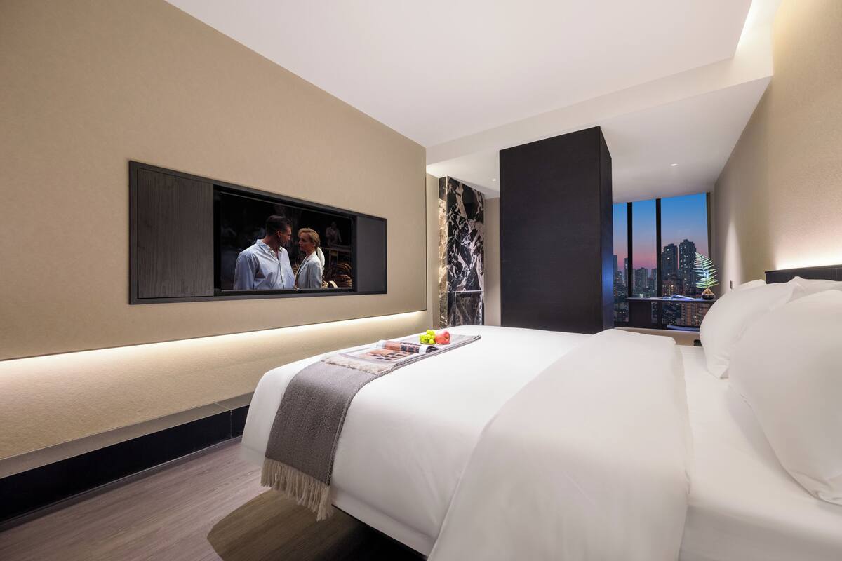 A modern hotel room with a large bed, a flat-screen TV mounted on the wall, and a cityscape view through the window.