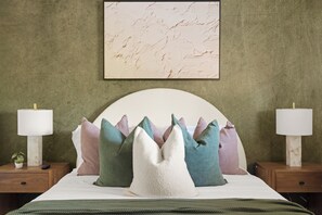 Una camera da letto con un letto, cuscini, un comodino, una lampada e un quadro appeso al muro.