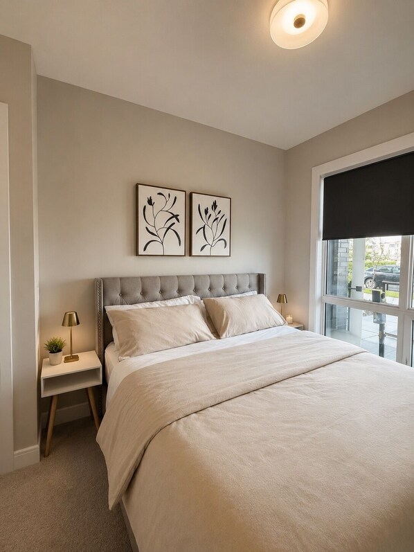 A bedroom with a bed, two nightstands, a window with a blind, and two wall-mounted artworks. - Currie Barracks Condo (Calgary)