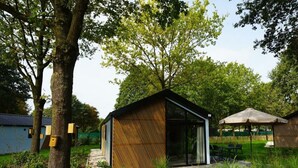 Una estructura moderna al aire libre con techo inclinado y grandes ventanales, rodeada de árboles y un césped.