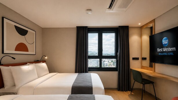 Ein Hotelzimmer mit zwei Betten, einem Schreibtisch, einem Sessel, einem Fernseher, der den Namen des Hotels zeigt, und einem Fenster mit Blick auf Gebäude.
