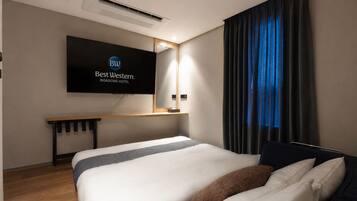 A hotel room with a bed, a TV mounted on the wall, a desk, and a window with curtains.