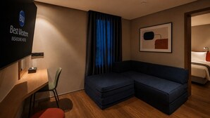 Ein modernes Hotelzimmer mit einem großen Bett, einem blauen Sofa, einem Schreibtisch mit Stuhl und einem Wand-Fernseher, der den Namen des Hotels zeigt.