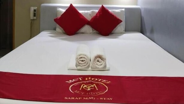 A hotel room with a bed, two red pillows, a red bedspread, and a towel arrangement.