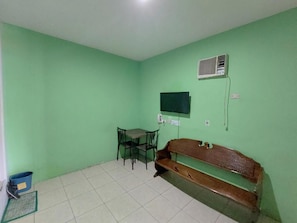 Une pièce aux murs verts, avec un banc en bois, une table et des chaises, et un téléviseur fixé au mur.