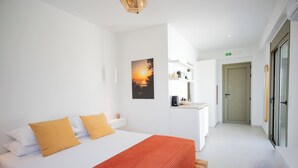 A modern hotel room with a bed, a red patterned throw, a painting of a sunset, a kitchenette, and a door leading to another room. - Hillside Villa with Private Pool & Panoramic Views | Plakias & Preveli (Asomatos)