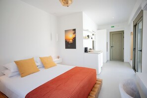 Ein modernes Hotelzimmer mit einem Bett, einer roten gestreiften Tagesdecke, einem Sonnenuntergangsbild, einer Küchenzeile und einer Tür zu einem weiteren Raum.