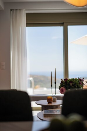 Ein Zimmer mit Blick durch ein großes Fenster, ein Tisch mit Kerze und Buch sowie eine Vase mit Blumen.
