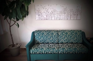 A teal sofa with a floral pattern, a potted plant, and a wall-mounted artwork depicting trees and birds. - A whole house near Palmanova, with a private entrance and garden. (Ontagnano)