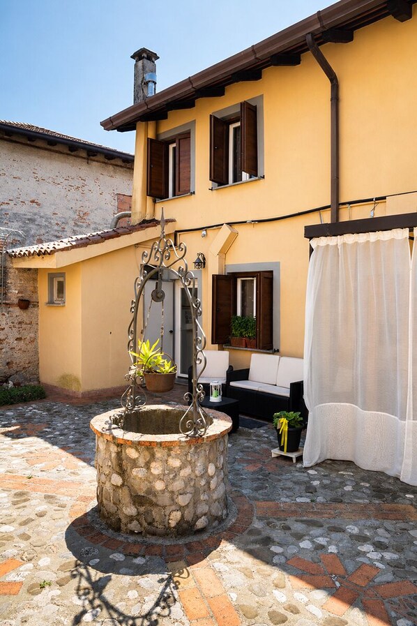 A courtyard with a stone well, outdoor seating, and a building with a yellow facade and brown shutters.