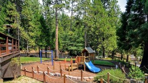 A playground with a slide, climbing structures, and a small building, surrounded by trees.